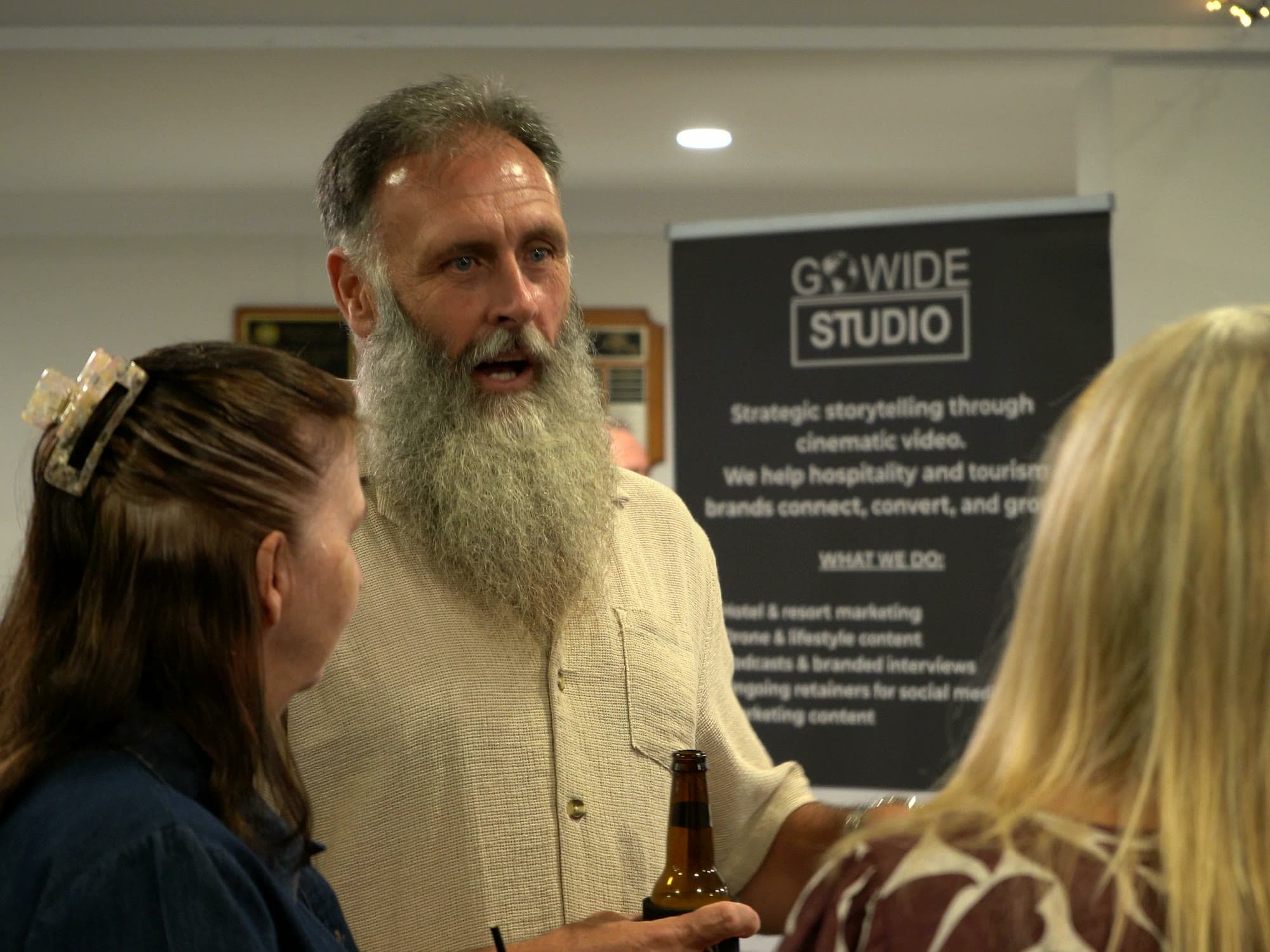 A bearded guest talking with two women beside the Go Wide Studio banner during networking.