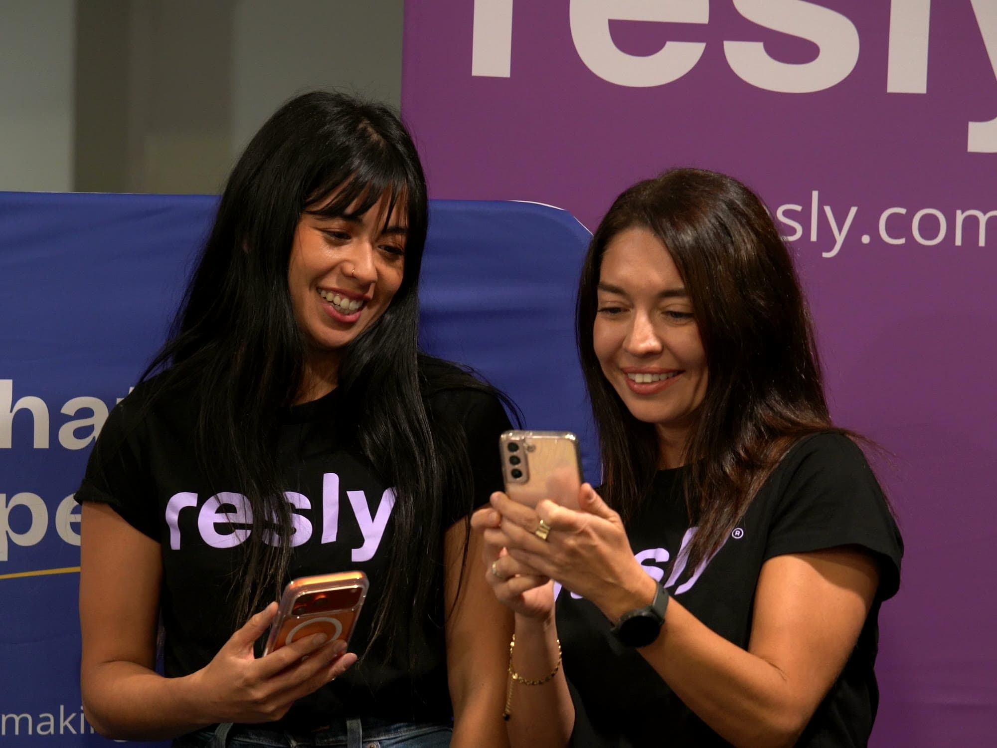Two members of the resly team smiling over their phones at the resly exhibitor stand.