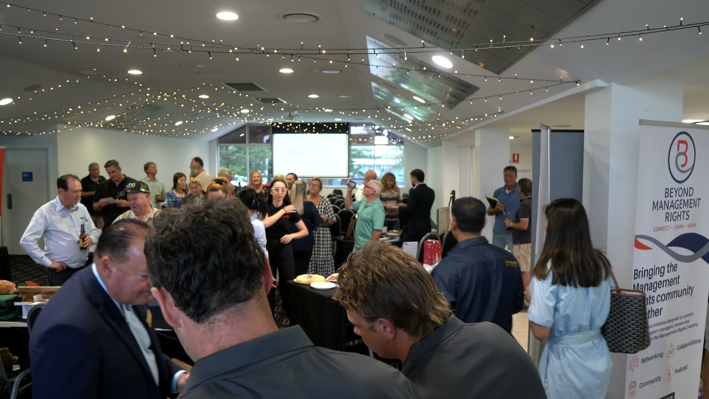 Guests networking in the function room at the inaugural Beyond Management Rights event, with festoon lights overhead and a Beyond Management Rights banner on the right.