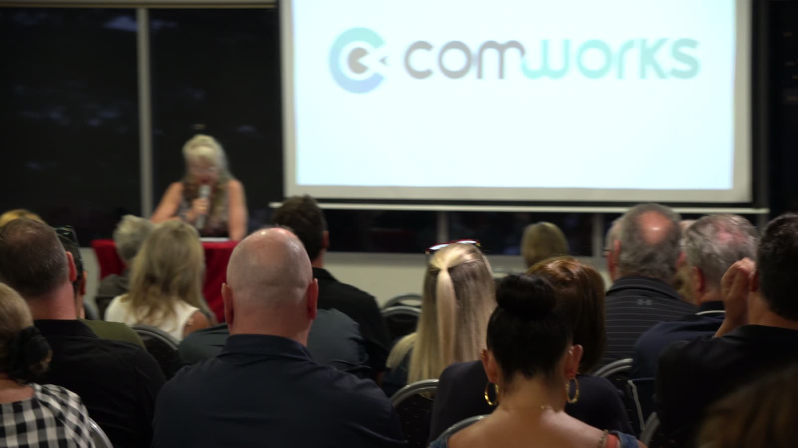 Attendees seated and watching a presentation, with an exhibitor logo on the screen behind.