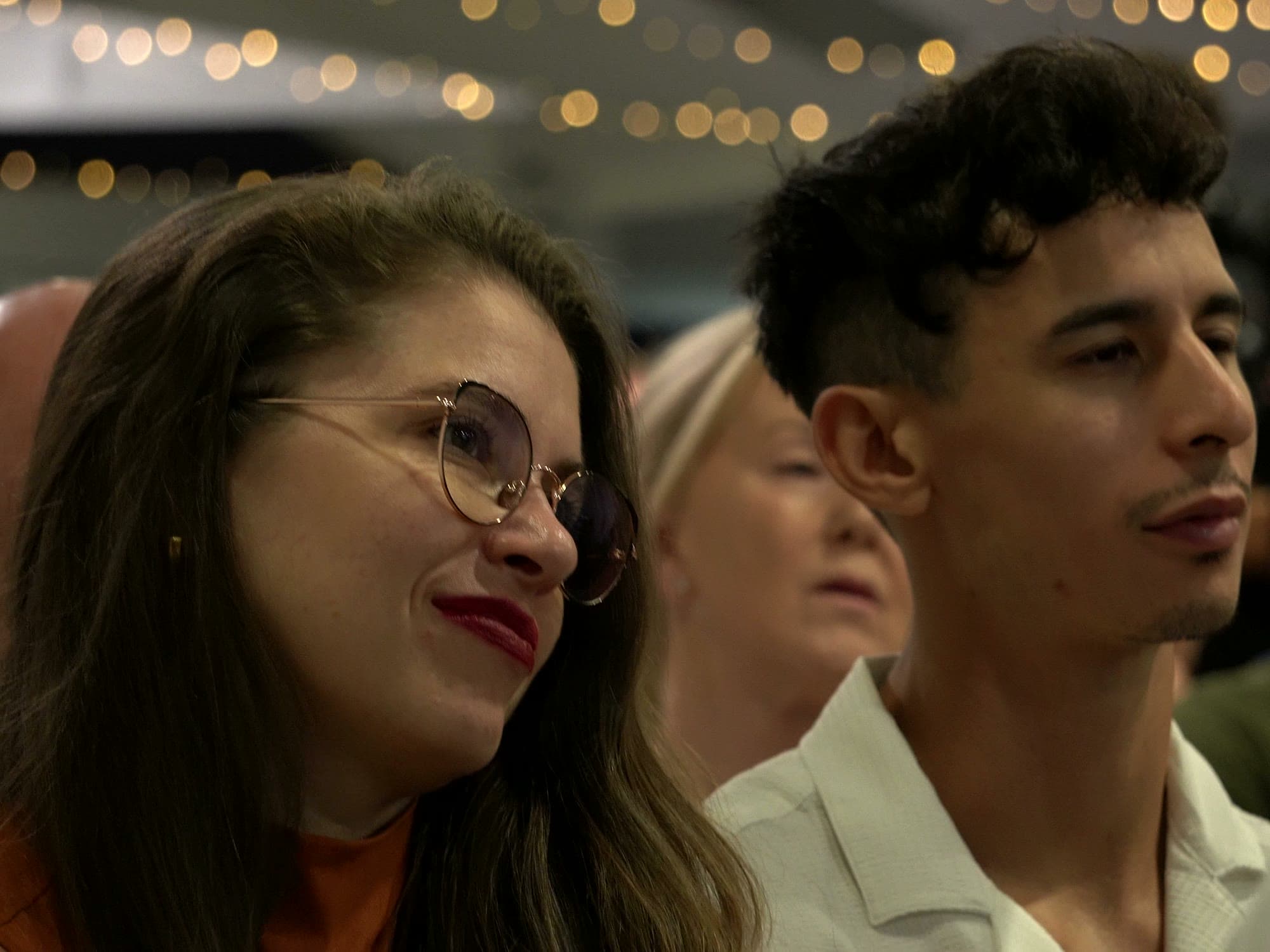 Two guests in the audience listening to the evening programme, one of them smiling, under festoon lights.