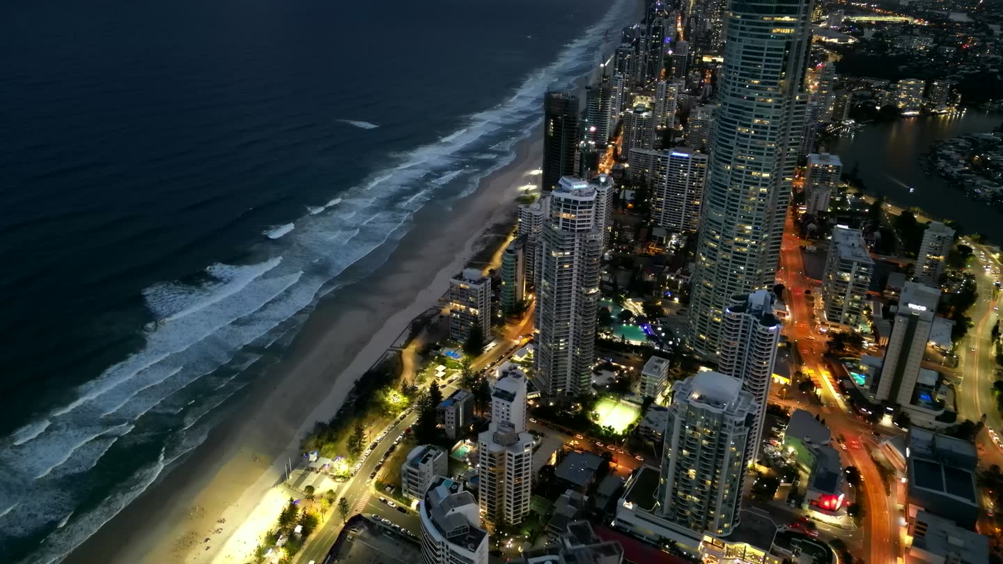 An aerial view along the Gold Coast coastline at dusk, with the beach on one side and lit accommodation towers on the other.